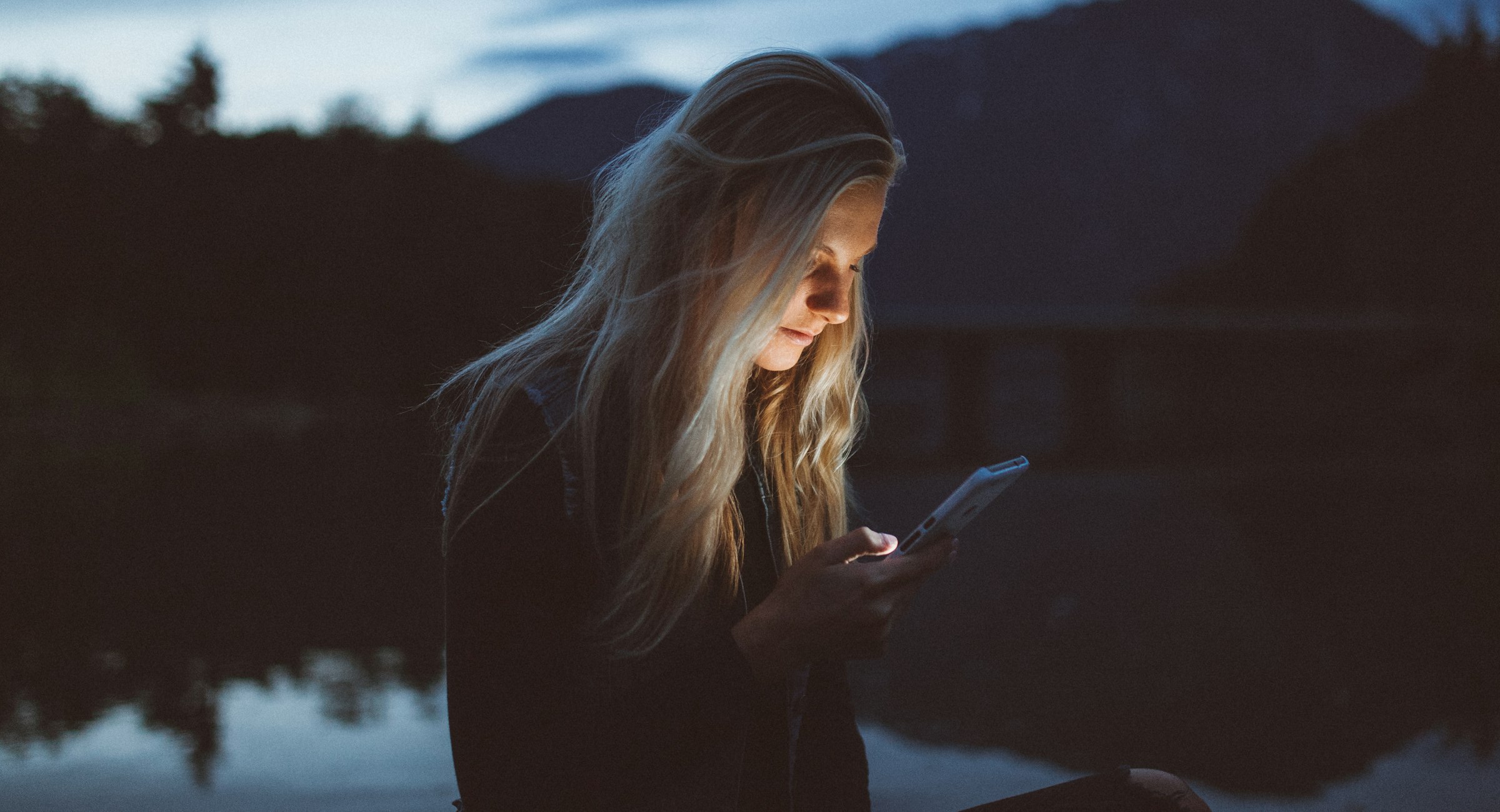 A person checks their phone by a lake at dusk